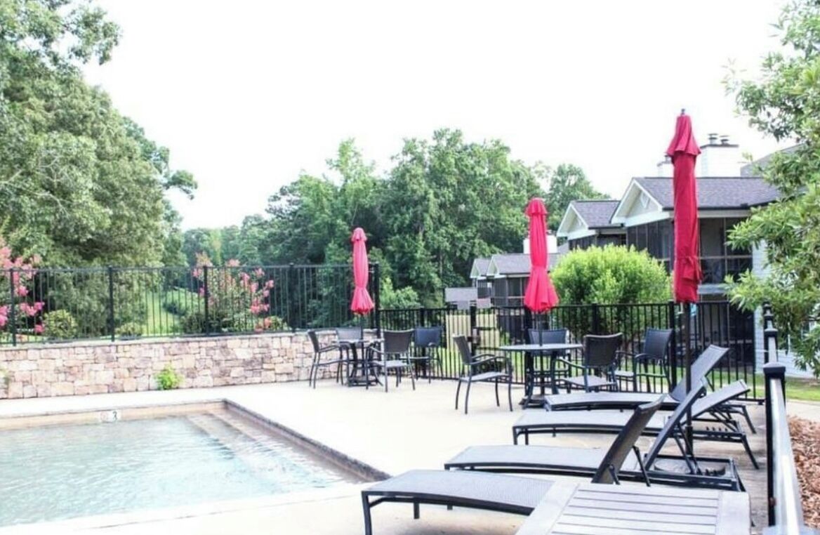 A poolside area with lounge chairs and red umbrellas, surrounded by a stone wall and greenery.