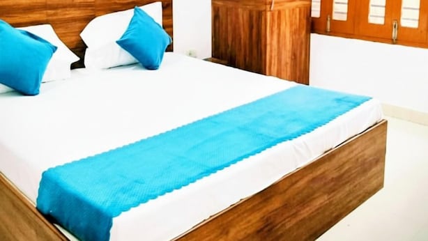 A bedroom with a wooden bed frame, white bedding, and blue pillows. There is a wardrobe with a mirror and a window with blinds. - Parvati Home Stay (Varanasi)