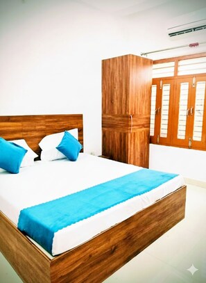 A bedroom with a wooden bed frame, white bedding, and blue pillows. There is a wardrobe with a mirror and a window with blinds. - Parvati Home Stay (Varanasi)