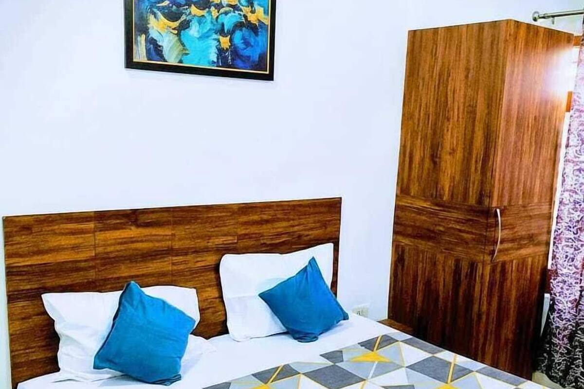 A bedroom with a wooden headboard, a bed with a geometric patterned bedspread, two blue pillows, and a wooden wardrobe.