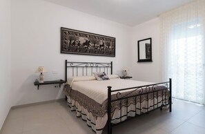 A bedroom with a bed, bedside tables, a wall-mounted tapestry, and a window with sheer curtains. - Domu S'Aratzia - 2km from the sea - with private parking (Quartu Sant'Elena)