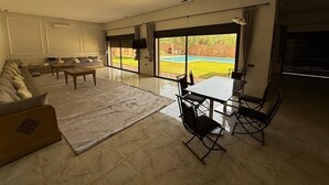 A spacious living room with a large sofa, a coffee table, and a dining area with a table and chairs. There is a sliding glass door leading to a pool.