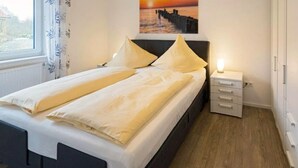 A neatly made bed with a white and beige duvet, a bedside table with a lamp, and a painting of a sunset over a pier on the wall. - Tillmanns Stadtlounge Exclusive 5 star vacation apartment in Norden. (Norden)