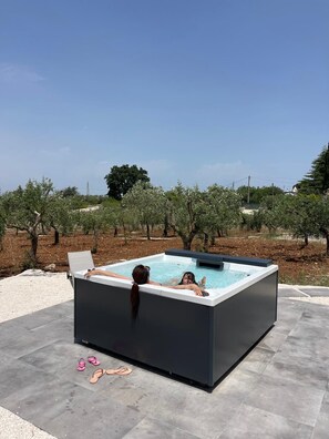 Tinaja exterior con dos personas relajándose, rodeada por un jardín ajardinado con árboles y cielo despejado.