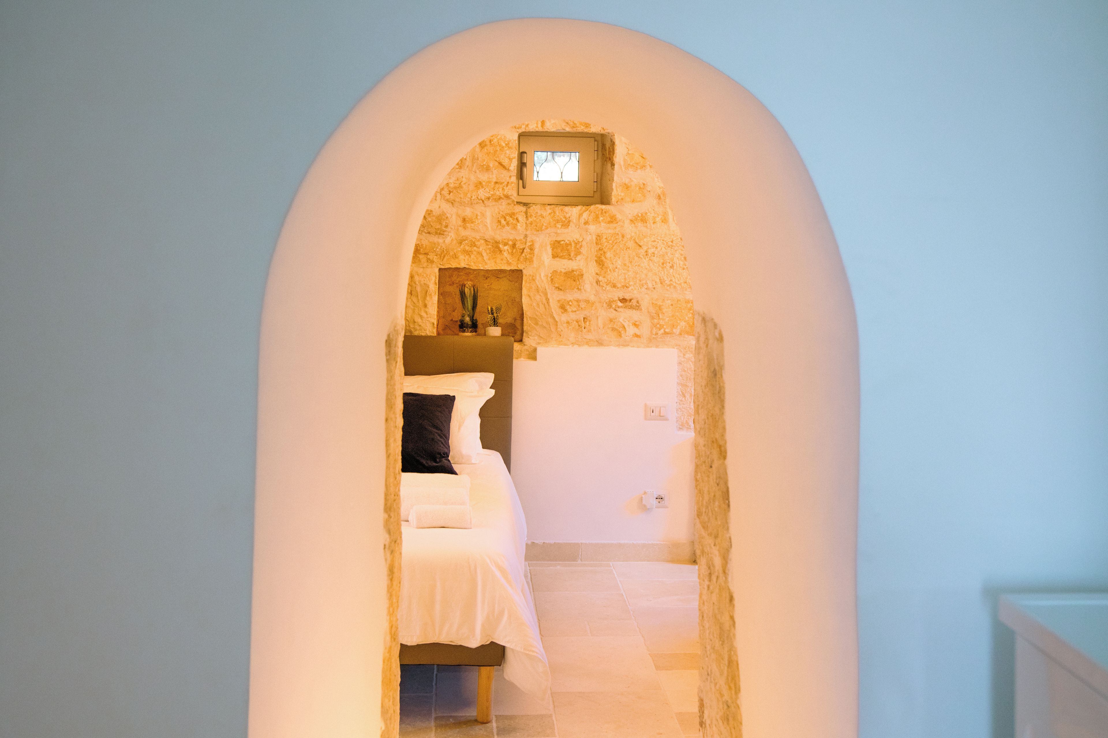A narrow hallway with a stone wall and an arched doorway leading to a bedroom with a bed and a nightstand.