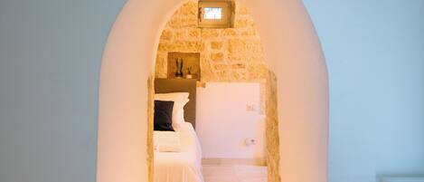 A narrow hallway with a stone wall and an arched doorway leading to a bedroom with a bed and a nightstand.