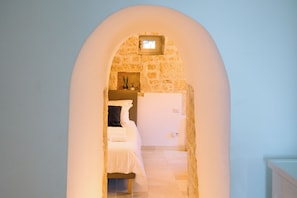 Un couloir étroit, avec un mur en pierre et une porte en arc, mène à une chambre à coucher qui contient un lit et une table de chevet.