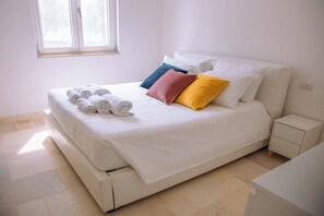 A neatly made bed with white linens and rolled towels, a bedside table, and a window with a view of trees.