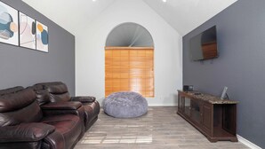 Una sala de estar con un sofá de cuero oscuro, un sillón de peluche redondo, un aparador de madera y un televisor de pantalla plana montado en la pared.