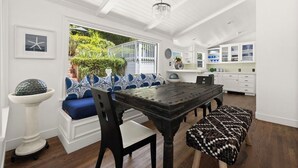 Un comedor con una mesa de madera, sillas y un banco. Se observa un pedestal blanco con un objeto decorativo. Fuera de la ventana, se ven una barandilla blanca y follaje.