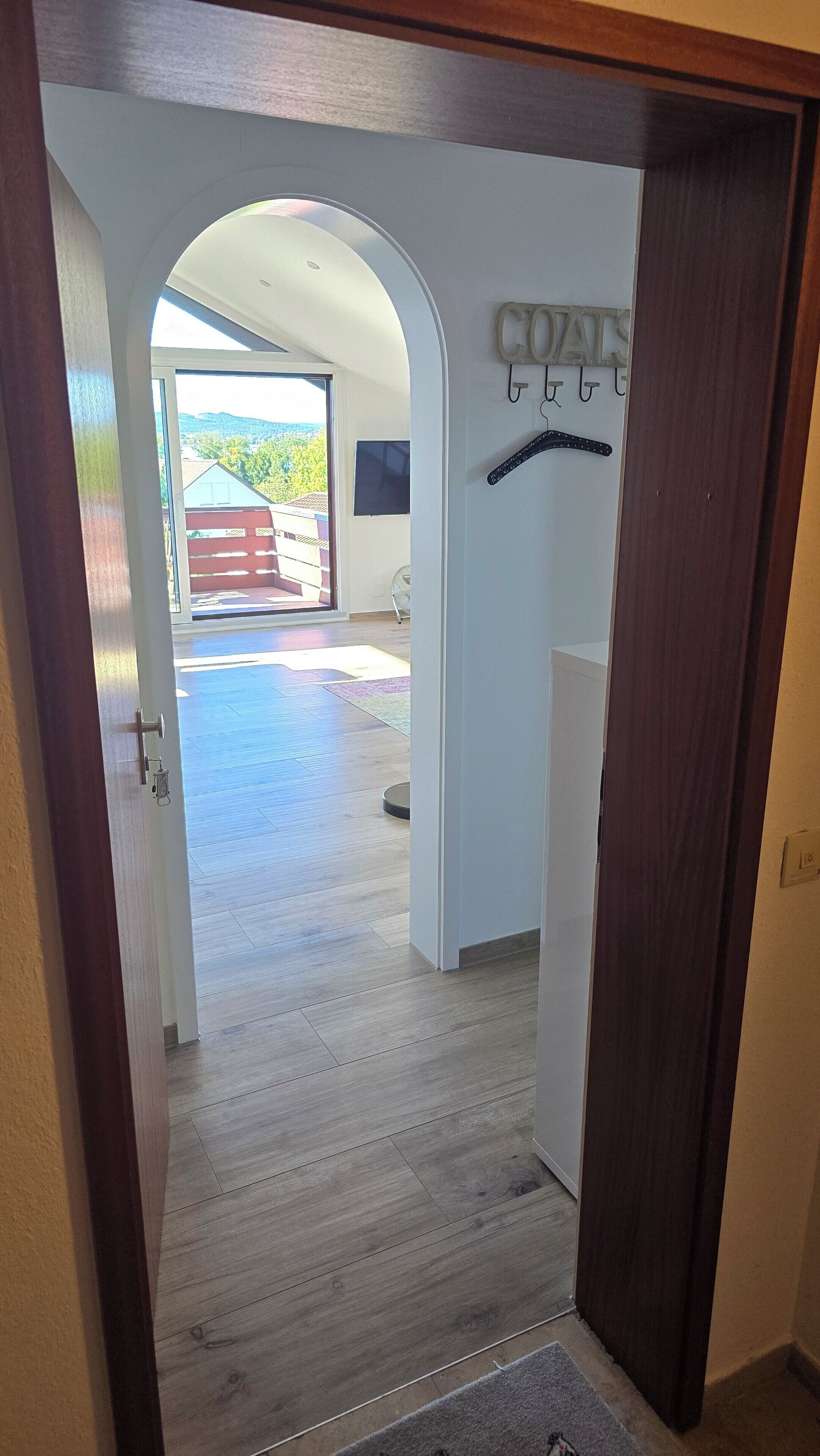 A doorway leading to a room with a wooden floor, a coat rack, and a view of a balcony with a red fence.
