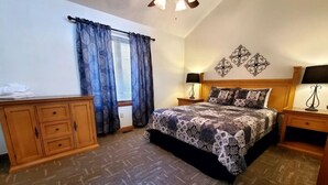 Une chambre à coucher comprenant un lit, deux tables de chevet, une commode en bois, un ventilateur de plafond et une fenêtre avec des rideaux.
