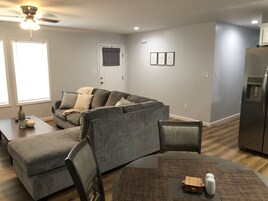 A modern living room with a grey sectional sofa, a dining table with chairs, a refrigerator, and a ceiling fan.