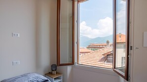 Une chambre avec une grande fenêtre, un lit, une table de chevet avec une lampe, et une vue sur des immeubles et des montagnes.