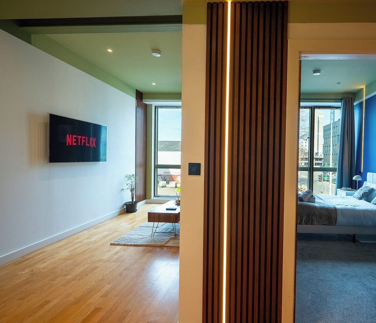 A modern living room with a flat-screen TV mounted on the wall, a wooden floor, a sofa, and a large window providing a view of the outside.