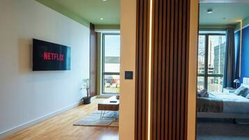 A modern living room with a flat-screen TV mounted on the wall, a wooden floor, a sofa, and a large window providing a view of the outside.
