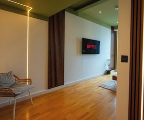 Una sala moderna con piso de madera, un sofá, un televisor montado en la pared mostrando 'NETFLIX' y un panel de madera vertical.