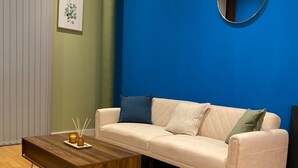 Ett vardagsrum med en beige soffa, ett soffbord i trä, en blå accentvägg, en rund spegel och en inramad tavla på väggen.