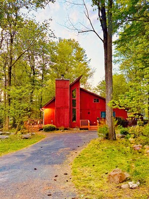 A red wooden cabin with a chimney, surrounded by trees and a gravel path. - Pocono Mountain CABIN-3 BDRS,HOT TUB, KING-BD, FIREPLACE, PET FRIENDLY,SLEEPS 8. (Tobyhanna)