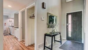 Un couloir doté d’un plancher en bois, une table noire sur laquelle se trouve un vase, un miroir encadré et une porte avec une fenêtre.