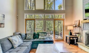 A modern living room with a sectional sofa, a fireplace, and large windows offering a view of trees. - Spacious 4BR Pocono Summit Home with Hot Tub (Pocono Summit)