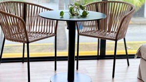 A round table with two wicker chairs, a potted plant, and a view of palm trees through large windows. - Modern Studio, Pool, WiFi, min Sea and 5th Ave. (Playa del Carmen)
