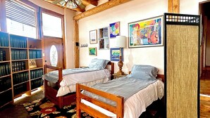 A room with two wooden beds, a bookshelf, a large window with blinds, and a ceiling fan.