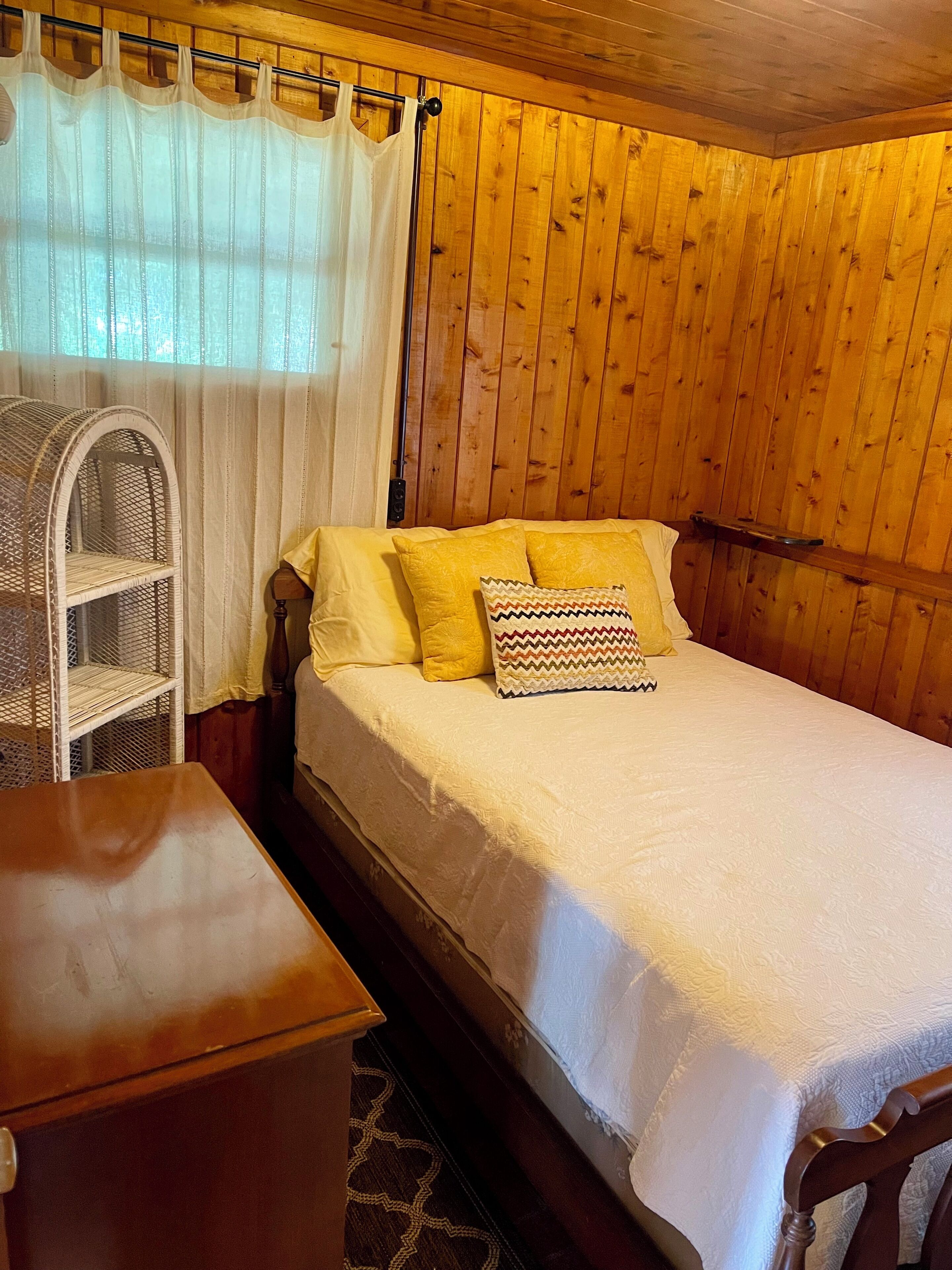 Une pièce aux murs lambrés, avec un lit, une chaise en osier et une petite table.
