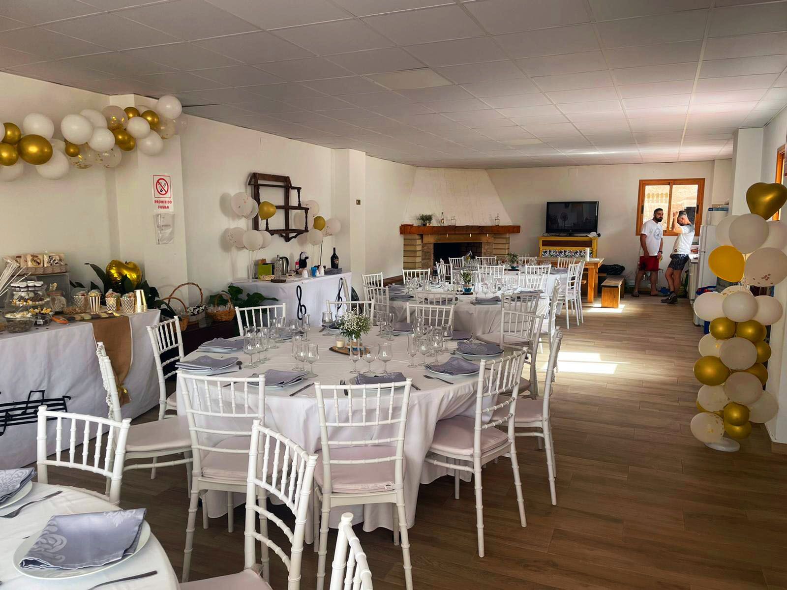 Un espace dédié aux repas, avec des tables rondes dressées pour un événement formel, des chaises blanches et une cheminée en arrière-plan.