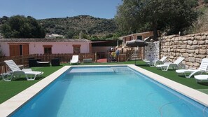 Una piscina rectangular con sillones blancos alrededor, una cerca de madera y un muro de piedra.