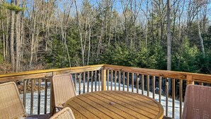 Une terrasse en bois, agrémentée de chaises en osier et d’une table ronde, entourée d’arbres et baignée d’un ciel dégagé.