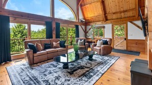 Ein geräumiges Wohnzimmer mit einer großen Holzbalkendecke, einer Ledersitzgruppe, einem Glastisch und Blick ins Freie durch große Fenster.