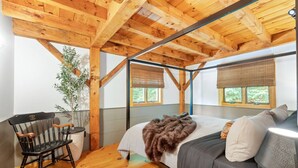Ein Schlafzimmer mit Holzboden, einem Bett mit grauer Bettwäsche, einem Stuhl und einem Fenster mit Blick auf Grünflächen.