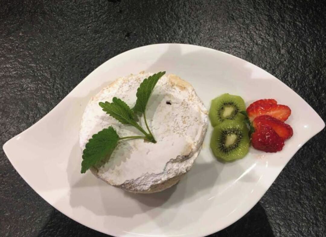 Un plat blanc amb un pastís cobert de crema i una fulla de menta, acompanyat de kiwi i freses en rodanxes.