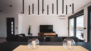Ein modernes Wohnzimmer mit einem schwarzen Sofa, einem Couchtisch mit Unterlage, einem Fernseher auf einem Holzgestell und einem großen Fenster mit Blick ins Freie.