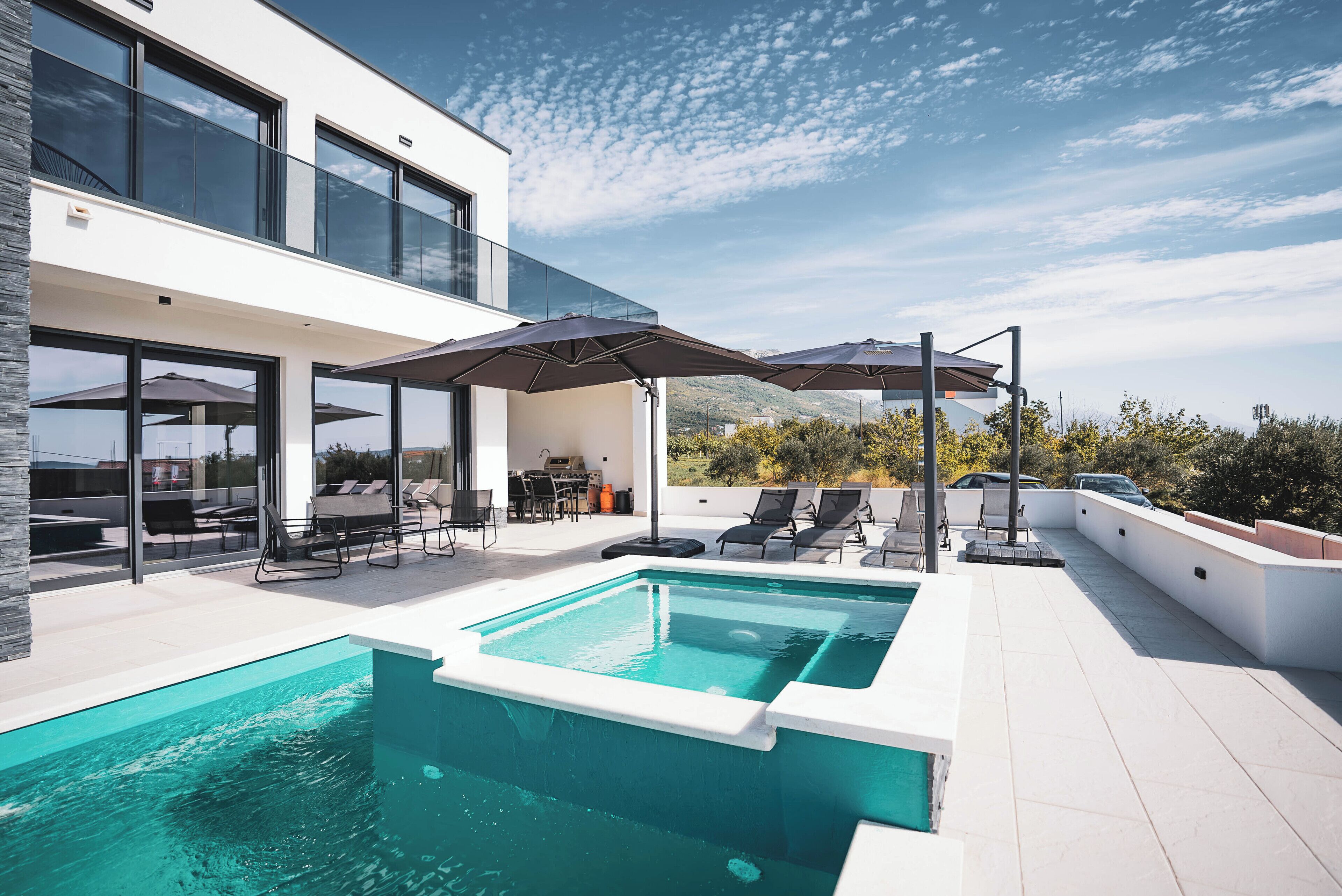 Ein moderner Poolbereich im Freien mit einer überdachten Terrasse, Liegestühlen und Sonnenschirmen.