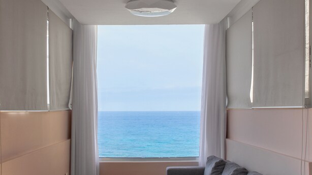 A room with a grey sofa, a window with curtains, and a view of the sea.