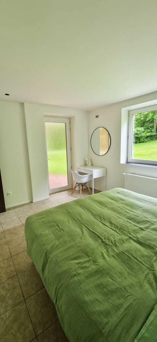 Une chambre à coucher comprenant un lit, un bureau, une chaise, un miroir et une fenêtre donnant sur un paysage verdoyant.