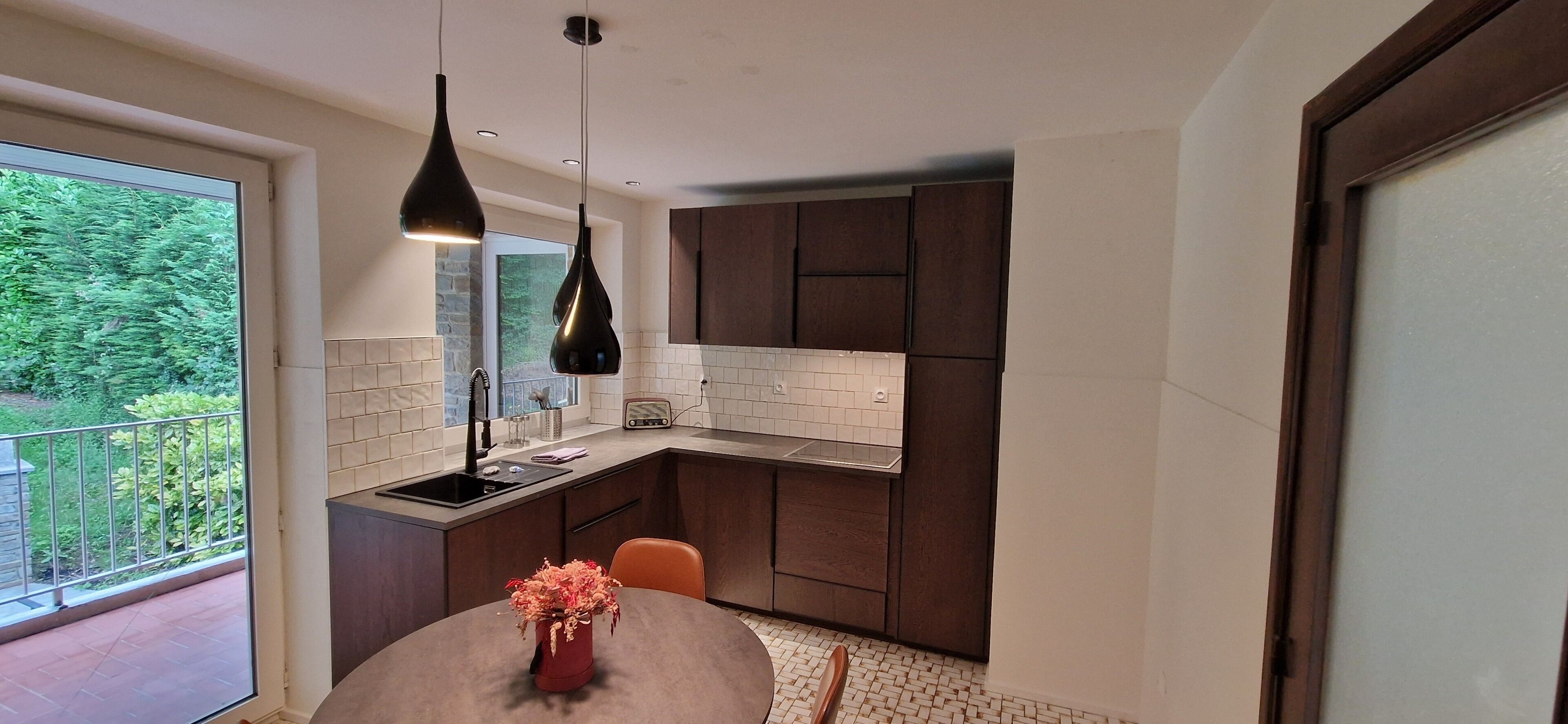 A modern kitchen with a built-in oven, a dining table with a vase of flowers, and a view of greenery outside.
