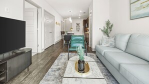 A modern living room with a flat-screen TV, a grey sofa, a coffee table with a decorative vase, and a dining area with a green chair. - BrightHaven Frisco - 1BR+ EV Parking near The Star (Frisco)