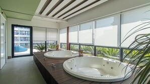 A modern bathroom with two large tubs, a wooden bench, and a view of a building and palm trees outside. - Loft a 10min do Metr com Academia na Vila Mariana (São Paulo)