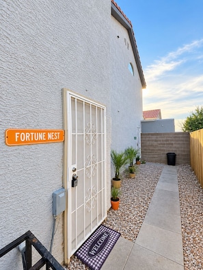Ein begrenzter Eingangsbereich mit einem Schild mit der Aufschrift 'FORTUNE NEST', einem Metallgitter, Blumentöpfen, einem Kiesweg und einer Fußmatte.