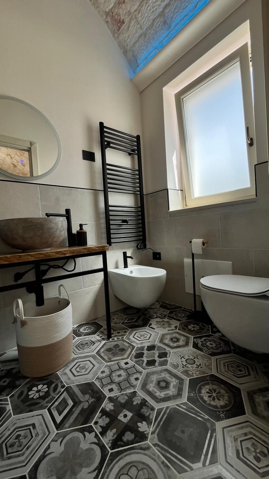 A bathroom with a large basin, a sink, and a toilet. There is a window above the toilet and a towel rack above the sink.