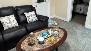 Un salon avec un canapé en cuir noir, une table basse agrémentée d’objets décoratifs, une porte blanche et une chambre à coucher visible à travers une porte ouverte.