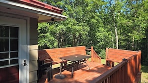 Una terraza con una mesa y bancos, rodeada por una valla de madera, y una casa con tejado rojo al fondo.