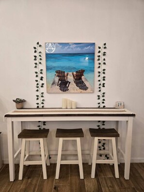 A white desk with three stools, a framed picture of a beach scene, and a small plant on the desk.