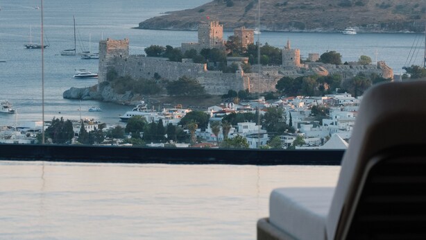 A view of a coastal town with historic buildings and a castle, viewed from a balcony with a wooden floor and a chair. - Mavilu Luxury Hotel (Bodrum)