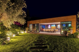 Un edificio moderno con una zona de estar exterior bien iluminada, fachada de vidrio y jardín ajardinado.