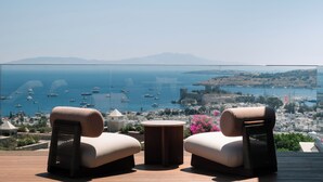 A balcony with two cushioned chairs and a small round table, overlooking a coastal town with boats and a clear view of the sea. - Mavilu Luxury Hotel (Bodrum)