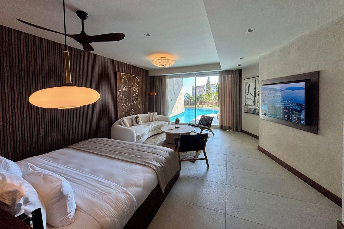 A modern hotel room with a bed, a sofa, a dining area, a ceiling fan, a TV, and a view of a swimming pool.